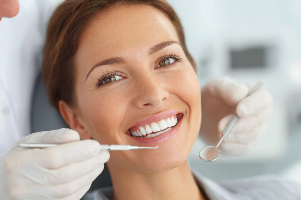 A young woman is smiling at the camera while receiving dental care, with a dentist using dental instruments and wearing white gloves in a modern, clean office setting.